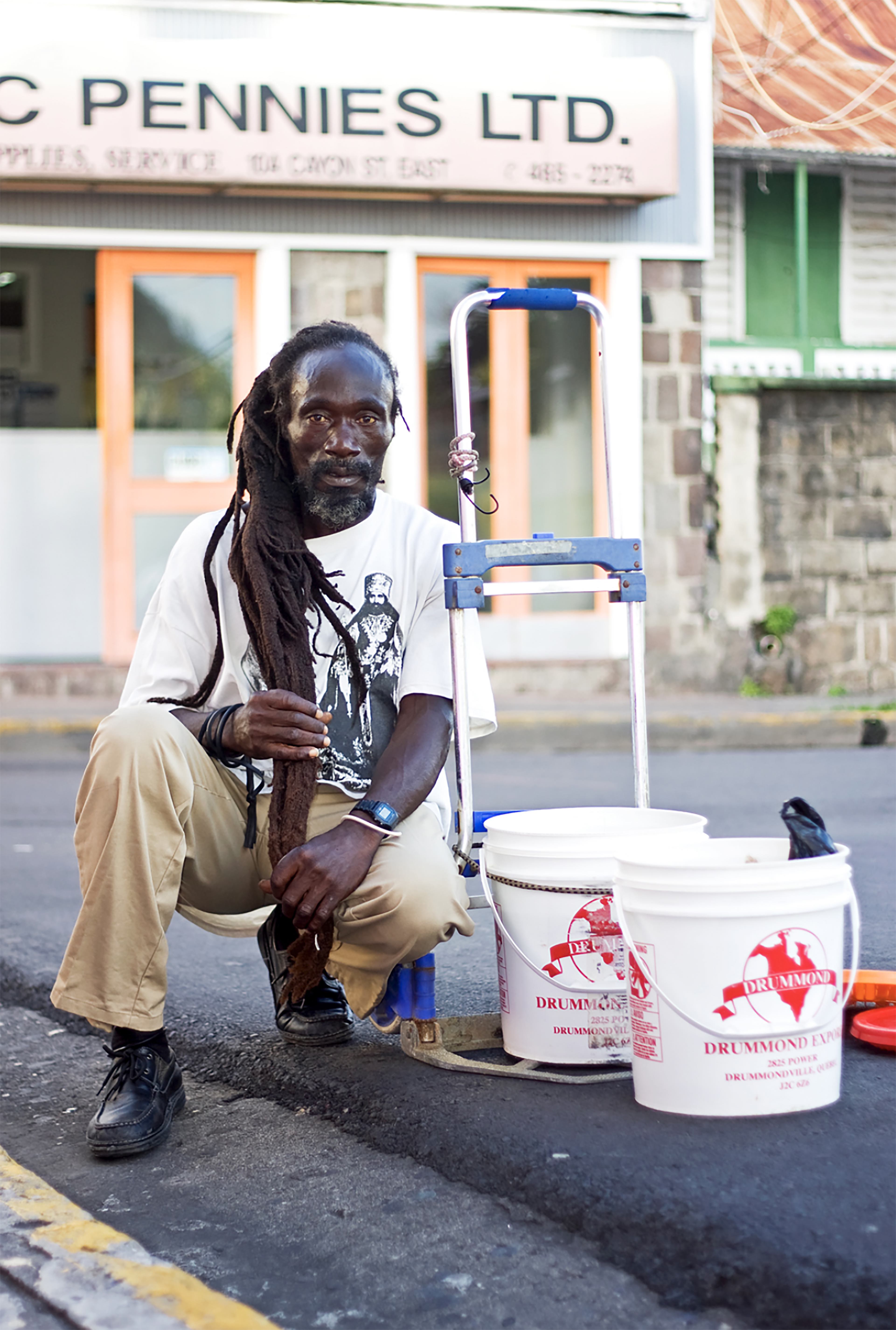 Dreadlocked man squatting next to handtruck and white plastic bins, on the curb.